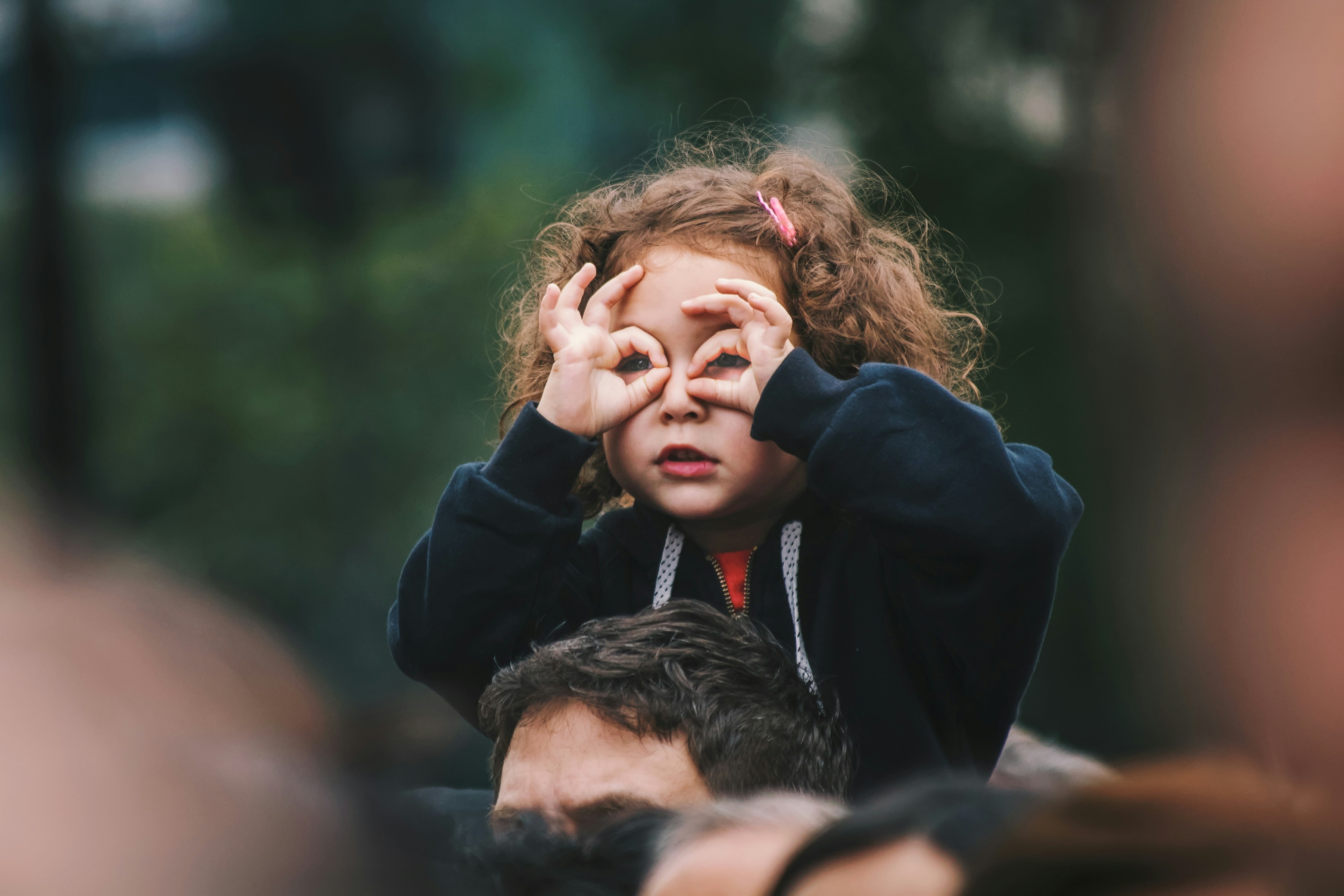 A child on adults shoulders using her fingers as binoculers